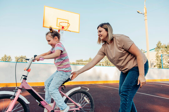 A young girl rides a pink bicycle while her mother supports her. They are enjoying quality time together in a sunny outdoor basketball court.
