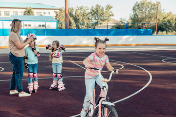 A family enjoys quality time at a sports court. A young Caucasian girl rides a pink bicycle. Two girls in roller skates play nearby, while a woman supervises them.