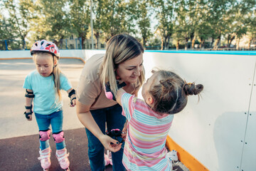 A woman interacts with two young girls at a skating rink. The woman has long brown hair and is smiling. One girl wears a pink helmet and the other has a striped shirt.