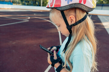 A young Caucasian girl with long blonde hair wears a colorful helmet and gloves while using a smartphone on a playground. The scene captures family bonding and quality time.