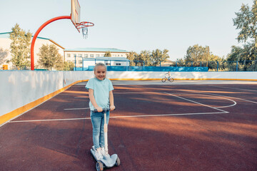 A young girl rides a scooter on an empty basketball court. She has short blond hair and wears a light blue shirt and jeans. The scene captures family bonding and outdoor activity.