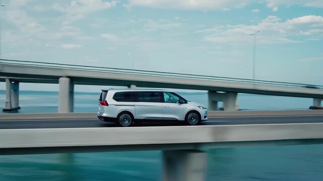 Silver minivan driving on a modern bridge over blue water under a cloudy sky vehicle