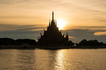 Chonburi, Thailand - November, 11, 2025 :Silhouette of an intricately carved wooden temple by the water at sunset with a golden sky and calm reflections