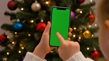 Child tapping green screen smartphone in front of a sparkling Christmas tree - Powered by Adobe