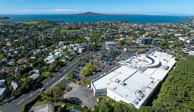 Aerial view of Eastridge shopping center and residential area in Auckland, New Zealand. Cars fill the parking lot as people shop. Rangitoto Island is visible in the distance.