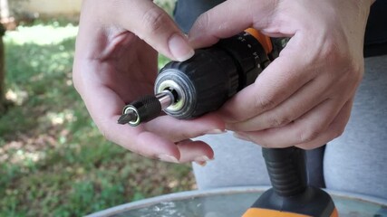 Close-up of female hands attaching a screwdriver bit to a cordless drill and testing it in an outdoor setting.