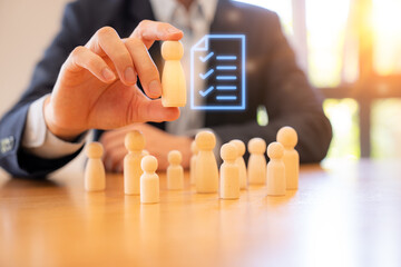 A business person selecting a wooden figure from a group, symbolizing human resources, recruitment, leadership, and employee evaluation. A digital checklist icon appears above the chosen figure.