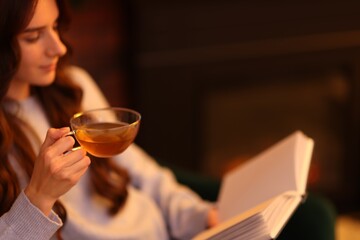 Woman with tea reading book near fireplace at home, selective focus