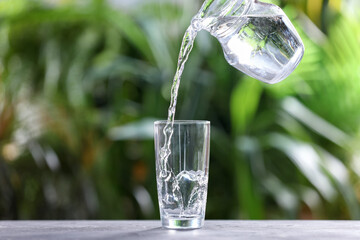 Pouring water into glass from jug at grey table against blurred green background, closeup. Space for text