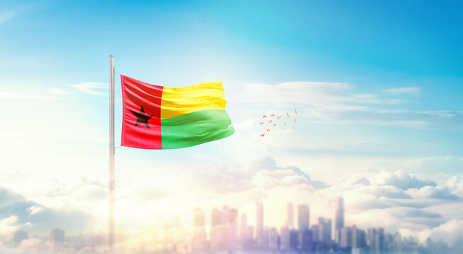 Guinea-Bissau flag wavering in beautiful clouds with building skyline. The flag with drone shot in beautiful sky. Guinea-Bissau national flag for independence day. - Powered by Adobe