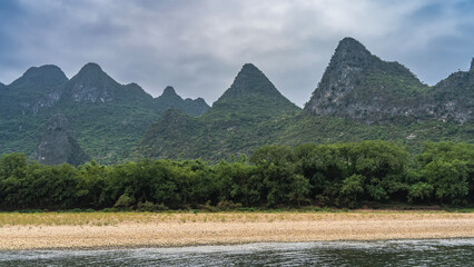 A picturesque river. Trees and bushes grow on the pebbly shore. Mountain range on the background of sky and clouds. China. Li River. Li Jiang. Guilin