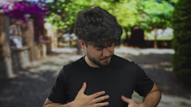 Young man with beard outdoors in a park, wearing black shirt, appears pensive with green foliage in background under bright natural sunlight.