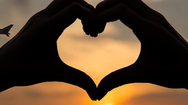 Silhouette hands forming heart shape with flying airplane against sunset