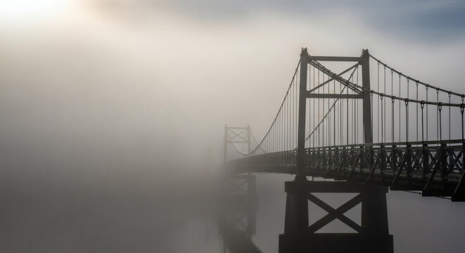 A solitary, antique suspension bridge disappearing into a dense, atmospheric morning fog over a river.