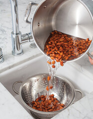 Pouring rinsed almonds from a metal pot into a metal strainer in a home sink. 