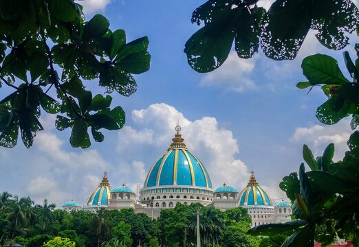 ISKCON the Indian Hindu Temple showcases its prominent blue and gold domes, set against a cloudy sky behind the green leaves and trees.