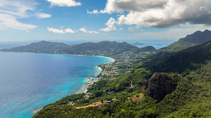 A turquoise bay along a coastal town surrounded by lush green hills under a partly cloudy sky. Seychelles, Mahe.