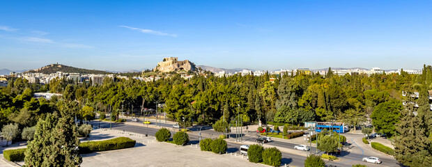 Bright daytime view of Acropolis hill above Athens, featuring busy avenue, green park trees and clear blue sky