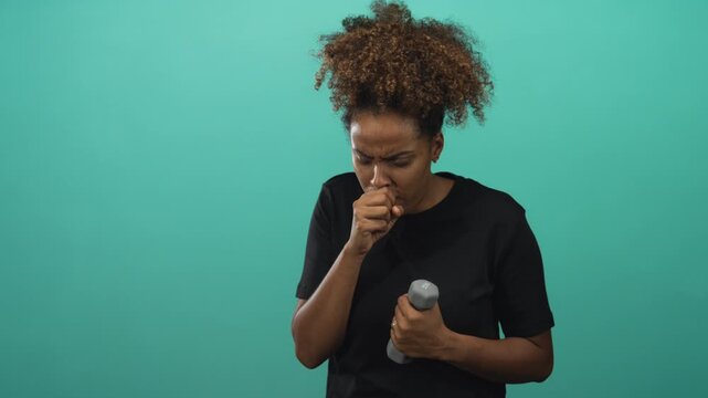 Woman young african american coughing into fist while holding grey dumbbell in teal studio setting with closed eyes and hand to mouth; health concern.