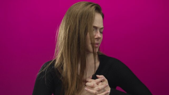 Woman clutch chest with interlaced fingers and serious expression in magenta studio; health risk alert awareness.