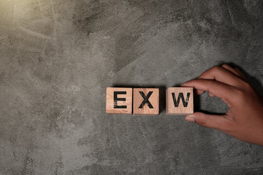 Wooden blocks displaying the letters &ldquo;EXW&rdquo; arranged on a textured gray surface, with a hand placing one of the blocks. Minimal business-themed layout suitable for logistics and trade concepts.