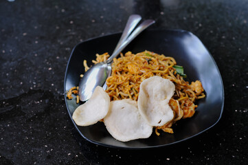 High-angle close-up of Indonesian Mie Goreng (fried noodles) served on a black plate with crackers and a spoon on a dark granite table.