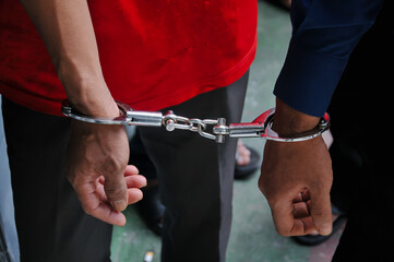 The wrists of a person wearing black and red clothing are bound together by heavy silver metal handcuffs, conveying themes of law enforcement, criminal apprehension, punishment, and legal proceedings.