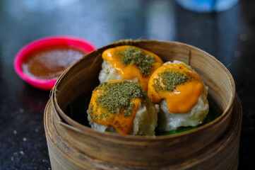 Three pieces of fresh, round dimsum or siomay dumplings, topped with orange cheese sauce and green dried seaweed seasoning, are presented in a classic bamboo steamer basket next to a bowl of pepper, c