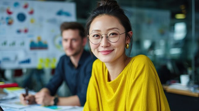 smiling asian businesswoman in yellow top looking at camera during a collaborative team meeting analyzing charts and financial data with a male colleague in a bright office - Powered by Adobe