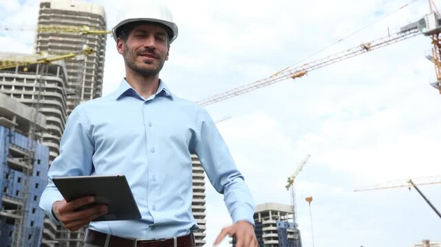 Side view of male engineer standing with digital tablet against background of buildings under construction working in construction helmet, points to the building and talks about the project - Powered by Adobe