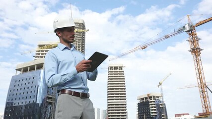 Side view of male engineer standing with digital tablet against background of buildings under construction working in construction helmet, points to the building and talks about the project