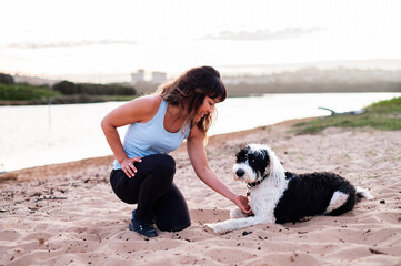 A woman kneels on the sand, engaging with her dog beside a calm river during sunset