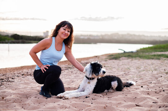 A woman kneels on the sand beside her black and white dog, smiling as the sun sets over the water