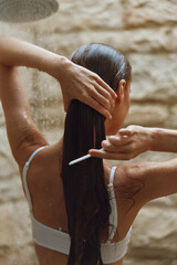 Woman combing long wet hair under shower with water drops, wearing white swimwear, natural stone background creating relaxing atmosphere and fresh summer vibe.