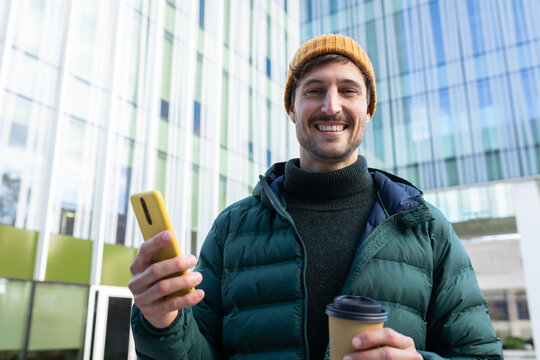 Smiling man holding coffee and using smartphone in urban setting - Powered by Adobe