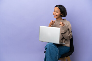 Young mixed race woman sitting on a chair with laptop isolated on purple background surprised and pointing front