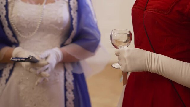 Women dressed in elegant vintage gowns and white gloves attending a formal event. One lady is holding a wine glass, and the other is holding a fan