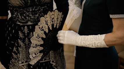 Two women wearing elegant historical dresses and white gloves holding vintage lace hand fans....