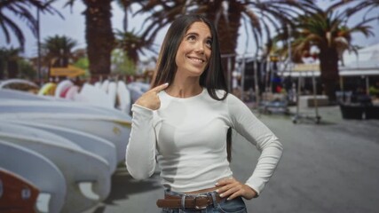 Young woman points finger to chest with surfboards and palm trees on a street; self belief confident.