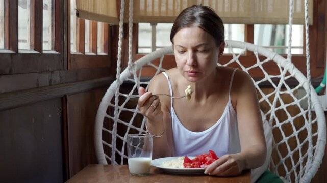 woman in a white tank top sits in a white rope hanging chair inside a dark wooden sunroom. She is eating a casual meal of fresh tomatoes and scrambled eggs from a white plate with a glass of milk.