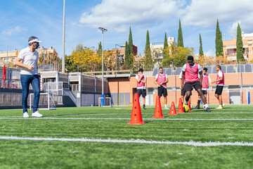 Football team training with a coach instructing players on a soccer field, dribbling around orange cones during practice