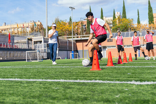 Young male football player practicing dribbling skills around cones, observed by a coach and teammates on a sunny sports field