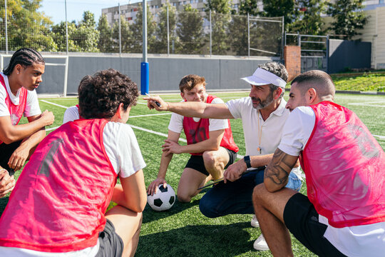 Soccer coach instructing young male players during training session, discussing game plan and tactics on a sunny field