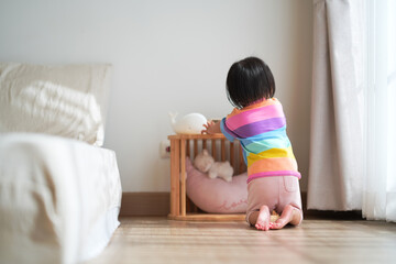 A young child wearing a colorful striped shirt kneels on the wooden floor in a cozy bedroom, looking toward a small wooden crib with soft toys.
