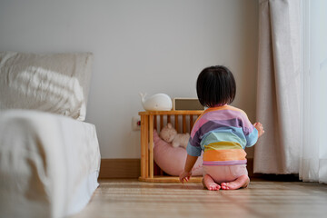 A young child wearing a colorful striped shirt kneels on the wooden floor in a cozy bedroom, looking toward a small wooden crib with soft toys.