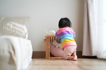 A young child wearing a colorful striped shirt kneels on the wooden floor in a cozy bedroom, looking toward a small wooden crib with soft toys.
