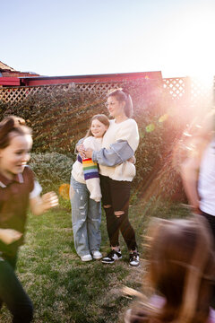 Mother hugging preteen daughter in the garden while young girls run around