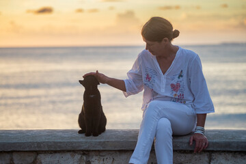 A woman in white sits on a stone pier by the sea next to a black cat