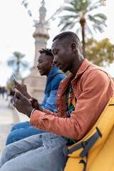 Obraz premium Two African men sitting outdoors, engrossed in their mobile devices, sharing a casual social media moment