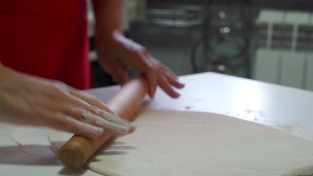 A thin sheet of white dough covers the floured table surface. Blurred hands energetically move a wooden rolling pin to flatten the pastry as the cook prepares a delicious homemade meal.